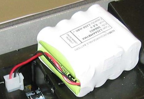 White battery pack with green cells and red wires, in battery compartment.